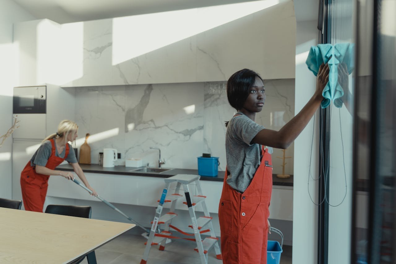 About Two women cleaning a modern kitchen, wiping glass surfaces and mopping the floor.